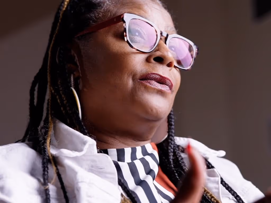 Close-up of a woman with braided hair wearing large glasses, hoop earrings, and a striped top under a white jacket.