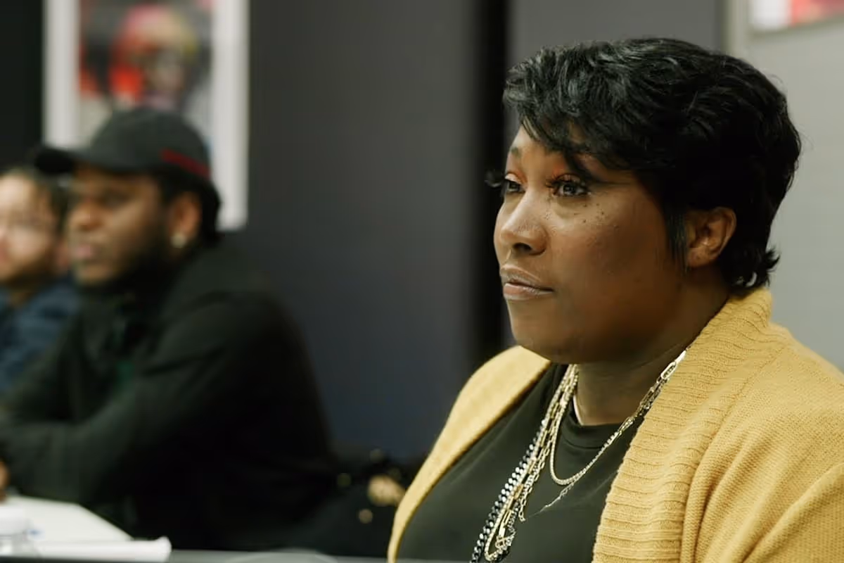A woman in a yellow cardigan attentively listens during a meeting with two blurred participants in the background.