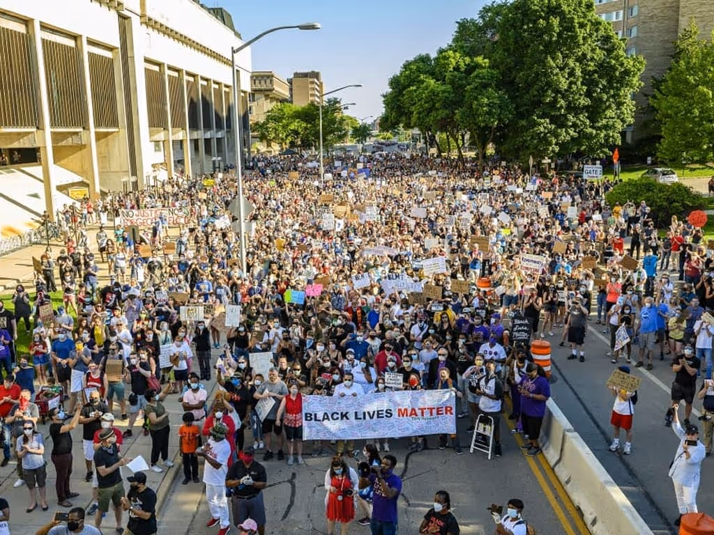 Large crowd of diverse protesters holding signs and a Black Lives Matter banner during a daytime march on a city street.