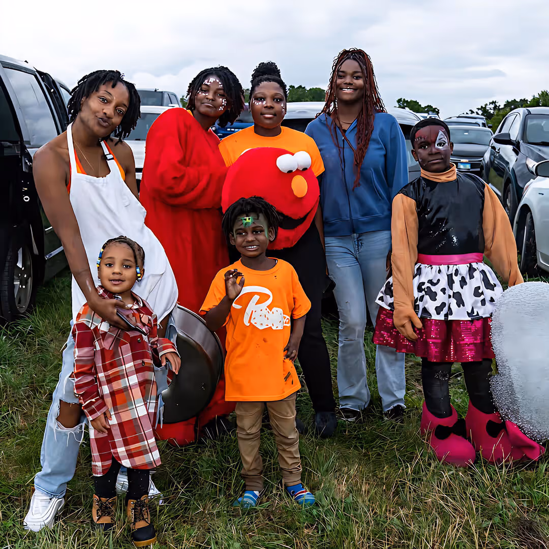 Group of children and adults with face paint and costumes standing on grass at an outdoor event with cars in the background.