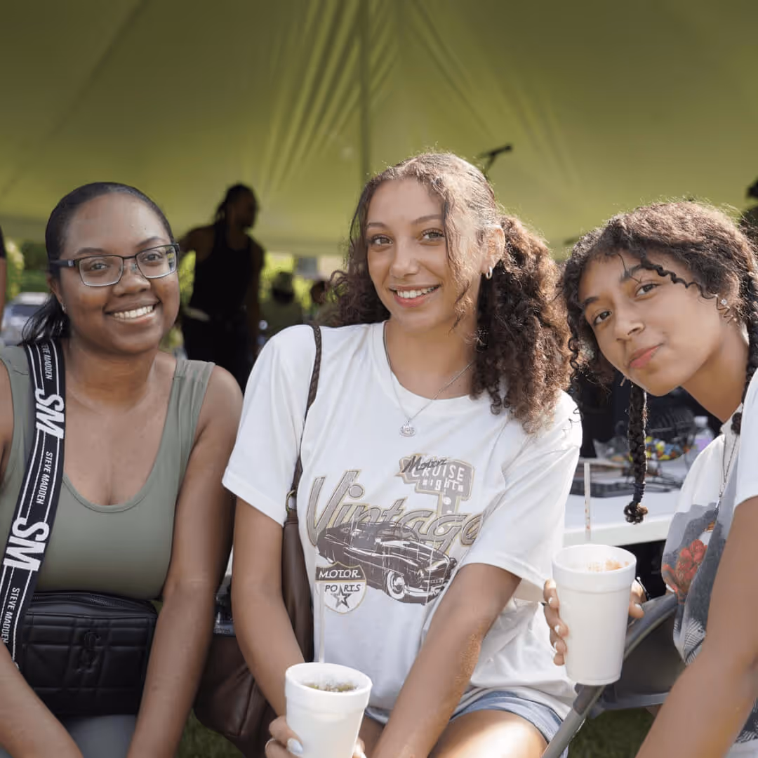 Three young women sitting outdoors under a tent, smiling and holding white cups.