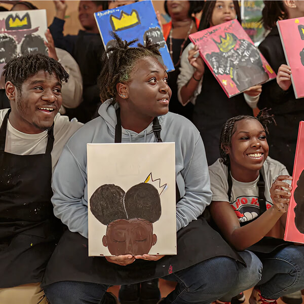 Youth holding painted portraits during a community art workshop, smiling and seated together.