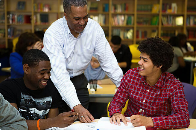 Adult mentor assisting two students with schoolwork at a table in a classroom or learning space.