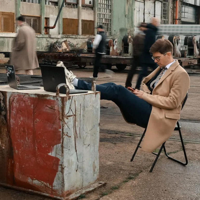 Two men working on laptops at a desk in a bright office with plants and large windows.