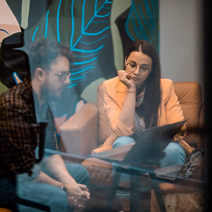 Two people sitting and discussing indoors with a laptop on a glass table.