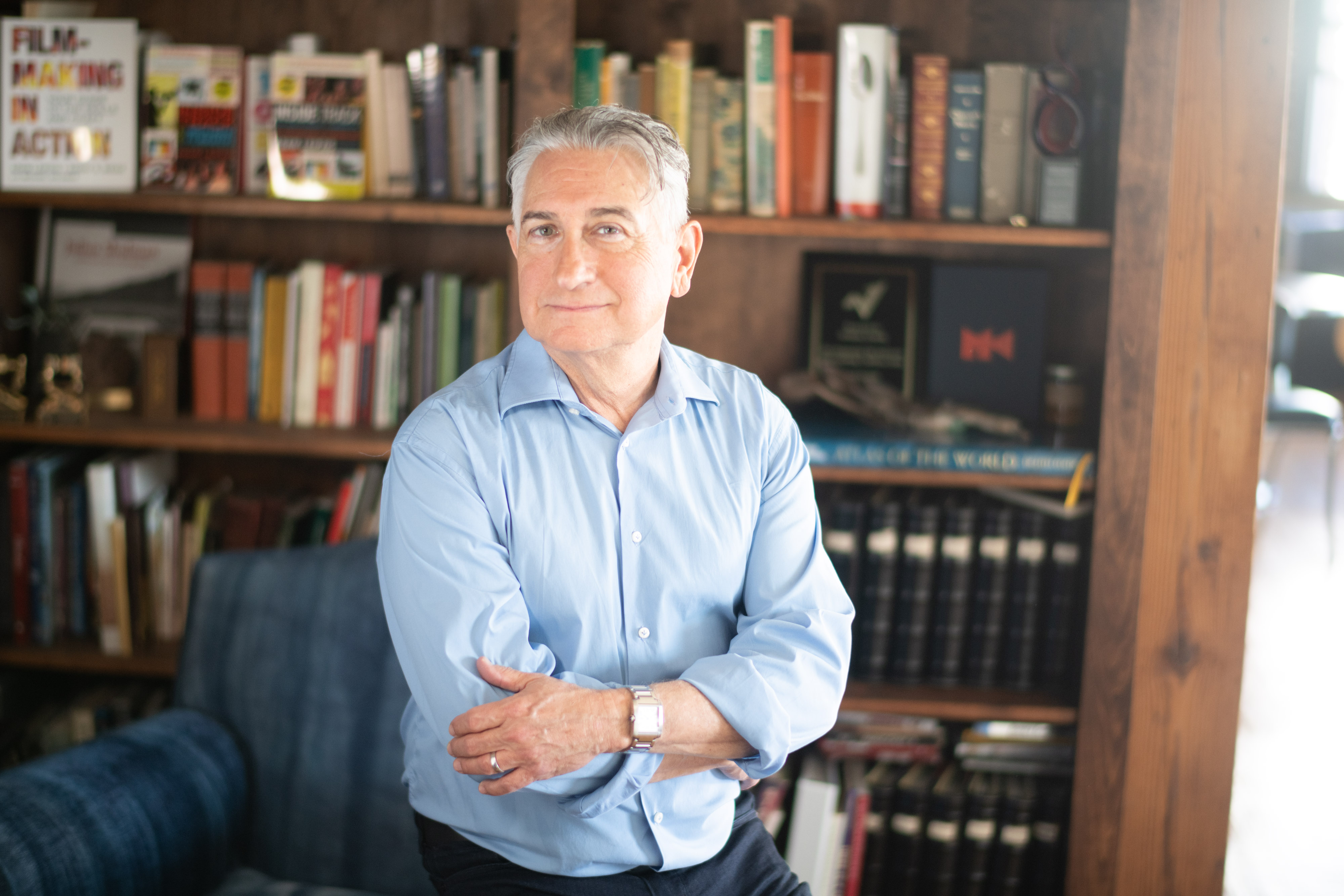 Adam Leipzig in a room with a shelf full of books in the background posing with his arms crossed