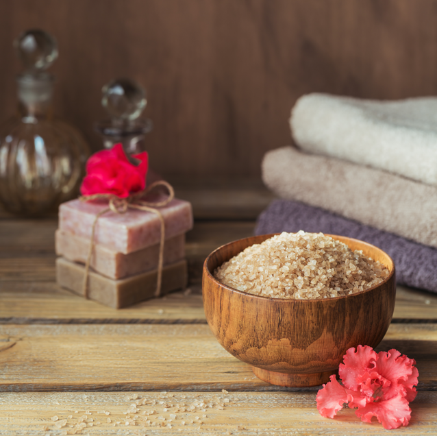Wooden bowl filled with bath salts, stacked soap bars tied with twine, folded towels, and a pink flower on a wooden surface.
