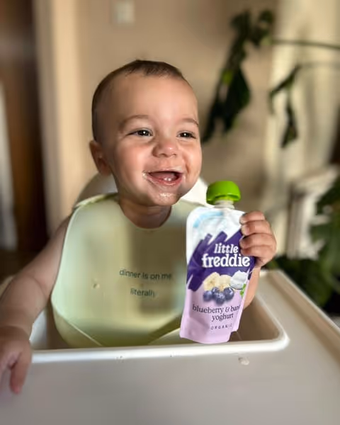 Smiling baby sitting in a high chair holding a pouch of Little Freddie blueberry and banana yoghurt.