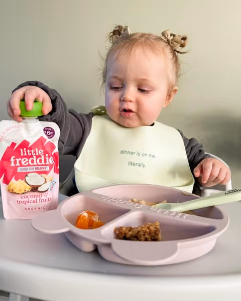 Toddler wearing a bib holding a pouch of Little Freddie baby food next to a divided plate with small portions of food.
