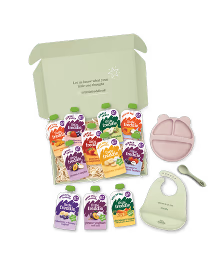 Open green box with assorted Little Freddie baby food pouches, a pink divided plate, a white bib, and a baby spoon.