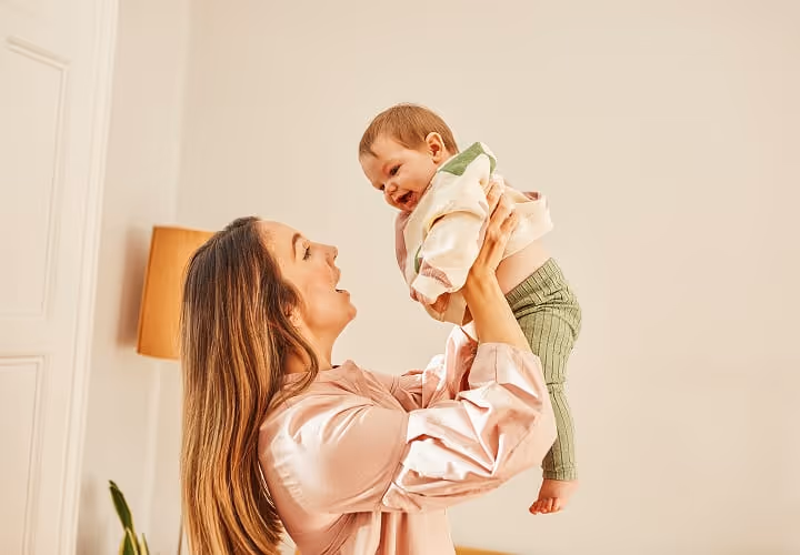 Woman lifting smiling baby indoors with beige wall and lamp in the background.