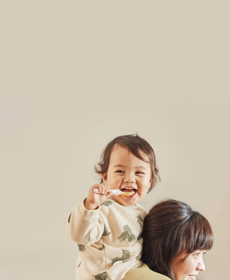 Smiling toddler with a spoon of Little Freddie food sitting on an adult's shoulders against a plain beige background.