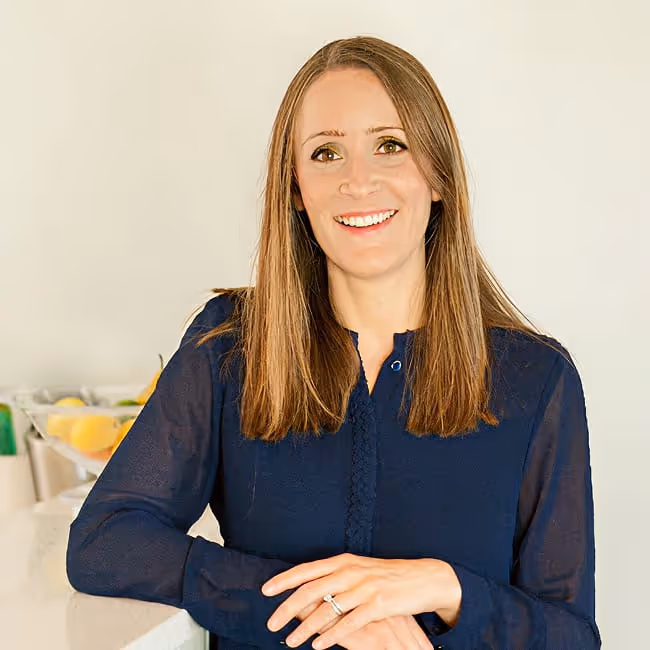 Jo Lenz, dietician, smiling with straight brown hair wearing a navy blue blouse, leaning on a white countertop in a bright room.