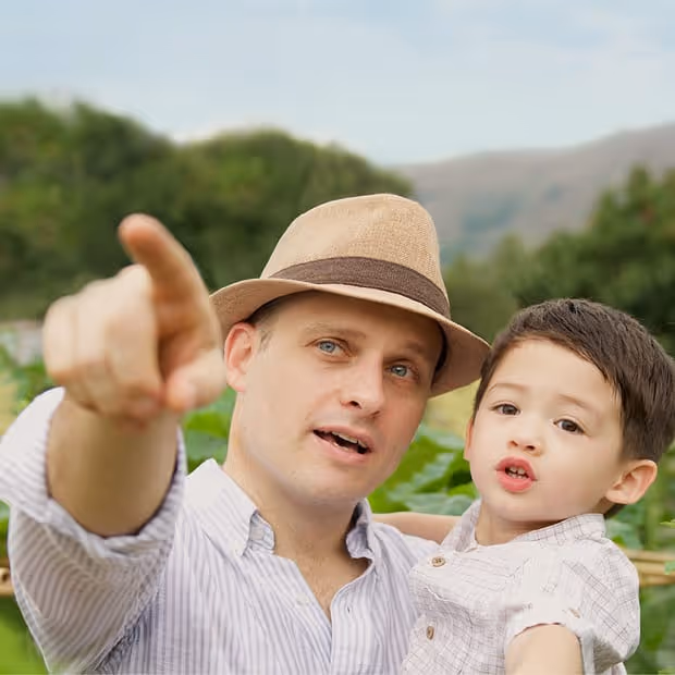Piers in a field pointing forward while holding a young boy outdoors with greenery and mountains in the background.