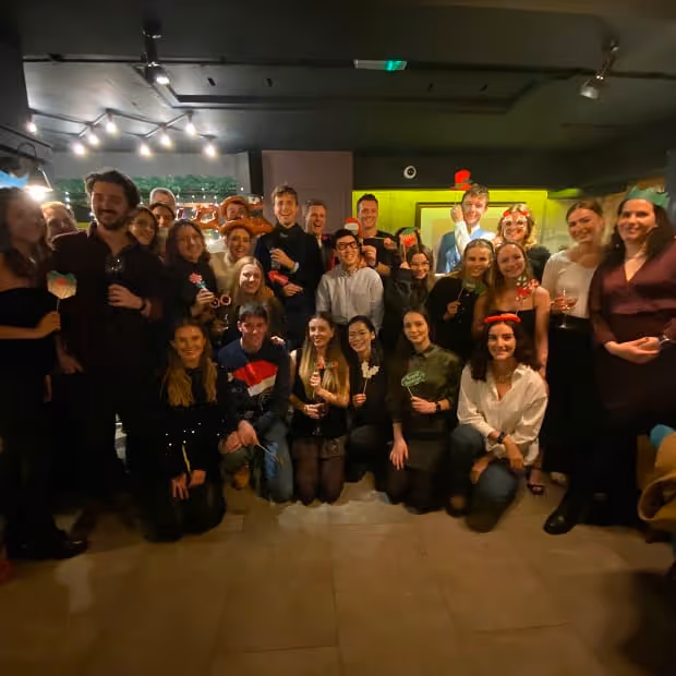 Group of Little Freddie employees posing closely together indoors, many holding drinks and small decorative props, smiling for a group photo under dim lighting.