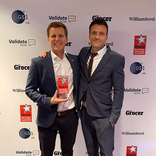 Two men in suits smiling and posing together, one holding a red Grocer New Product & Packaging Awards 2024 trophy.
