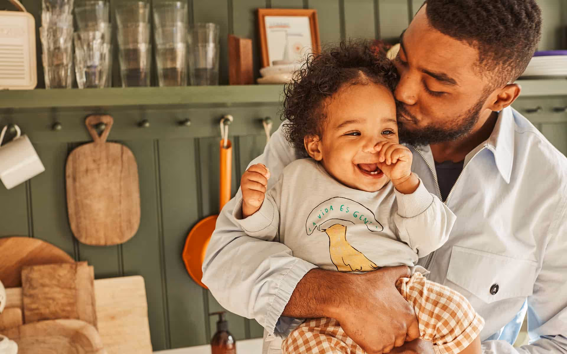 Man holding and kissing a smiling toddler in a kitchen with green cabinets and wooden cutting boards.