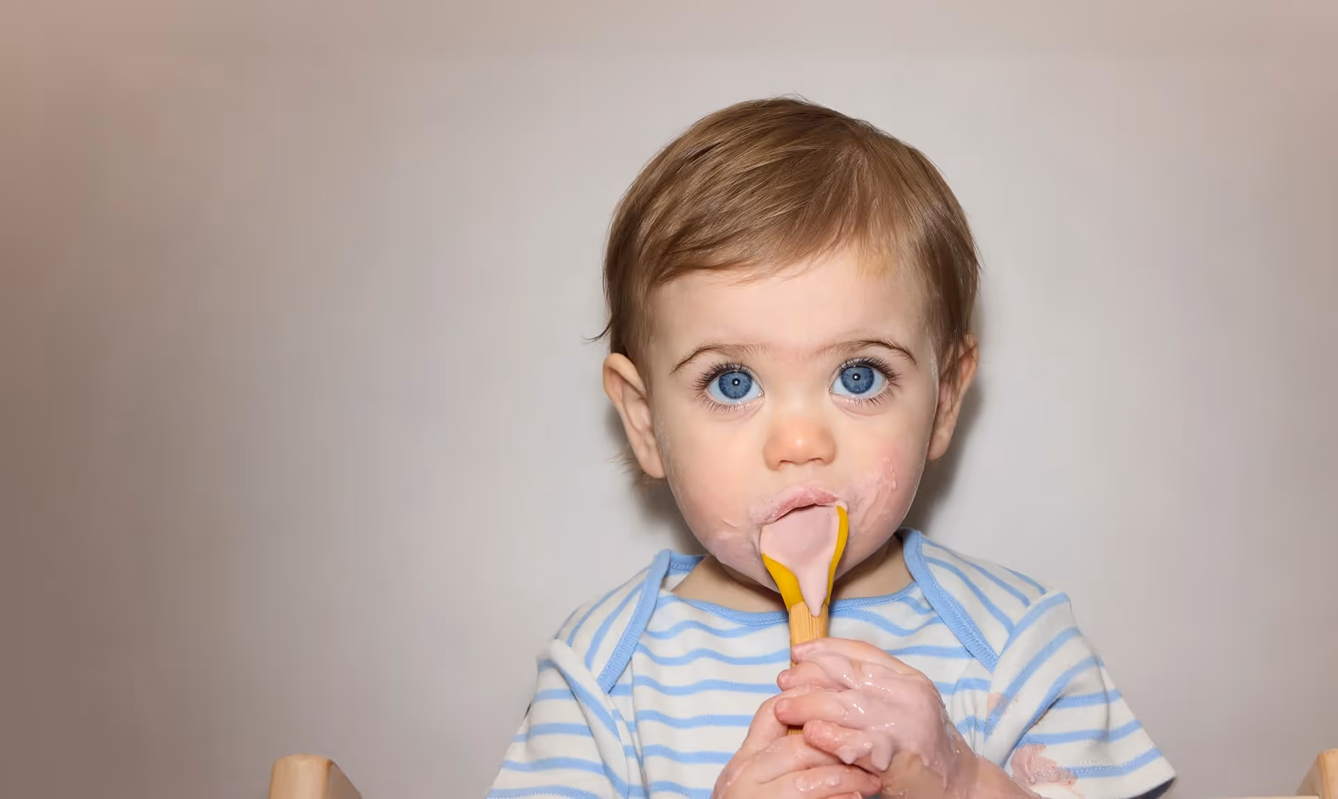 Toddler with blue eyes in a striped shirt licking a pink spoon with Little Freddie food smeared on face and hands.