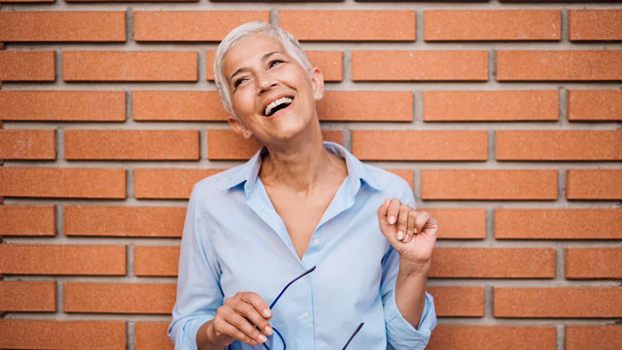 Smiling elderly woman with short grey hair wearing a light blue shirt holding glasses in front of a brick wall.