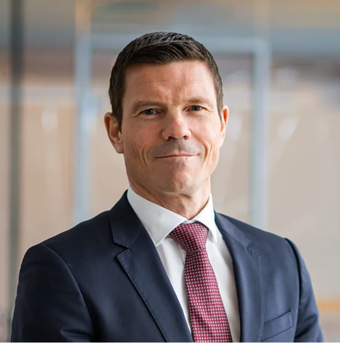 Portrett av en mann i dress og slips med kort mørkt hår som smiler svakt.