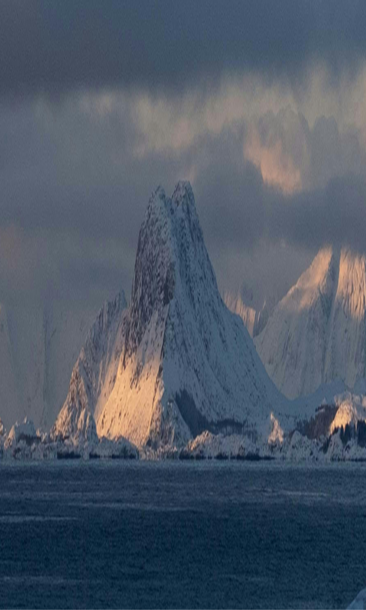 Snødekte fjelltopper med sollys som skinner på sidene ved en rolig sjø under en overskyet himmel.