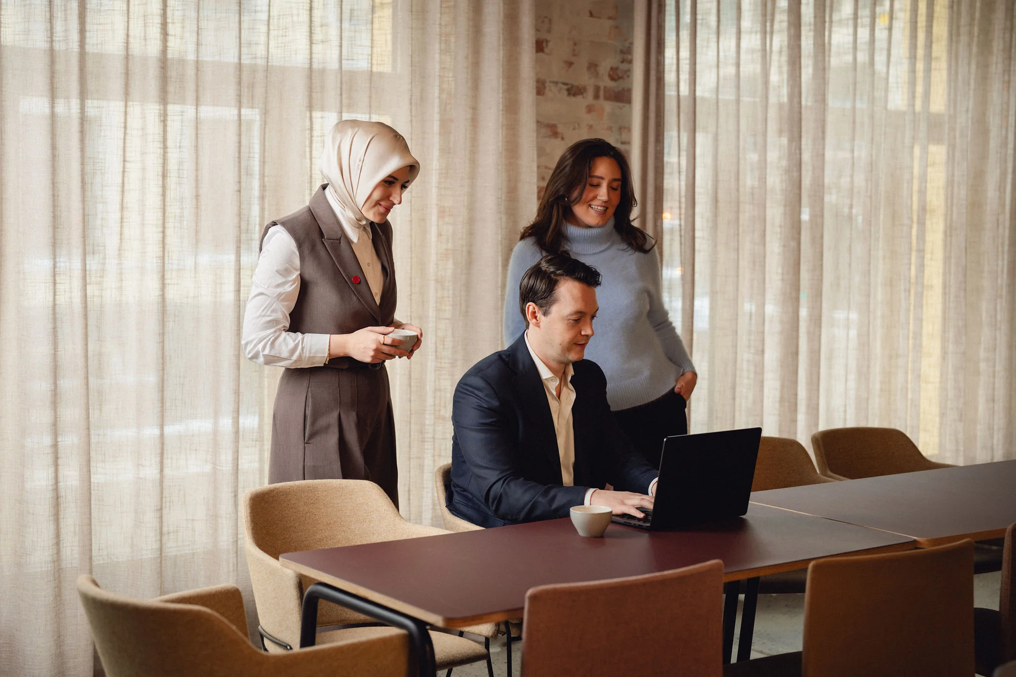 Tre personer, to kvinner og en mann, jobber sammen rundt et bord med en laptop i et lyst rom med lange gardiner.