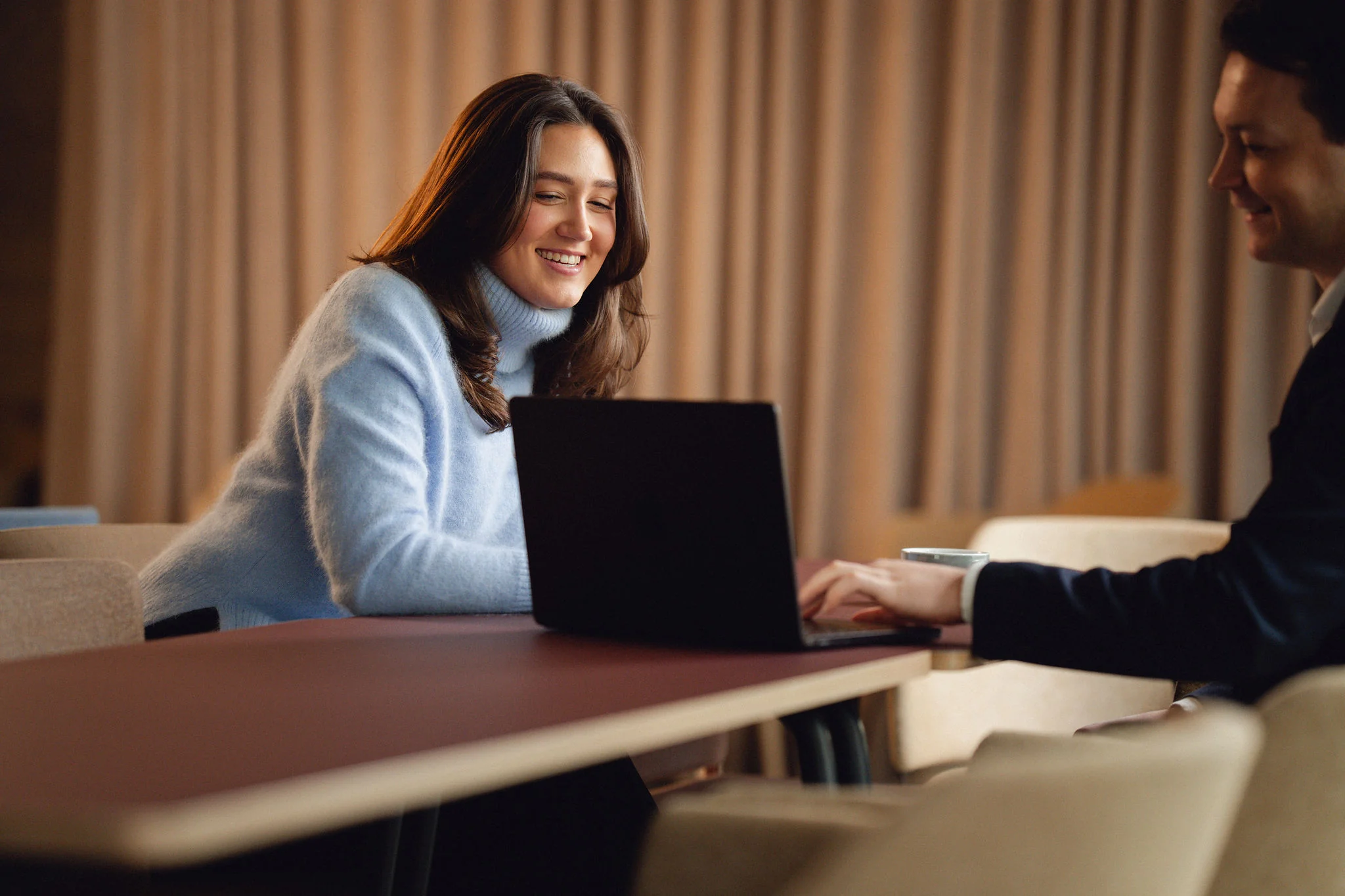 To smilende personer sitter ved et bord og ser på en bærbar datamaskin sammen.