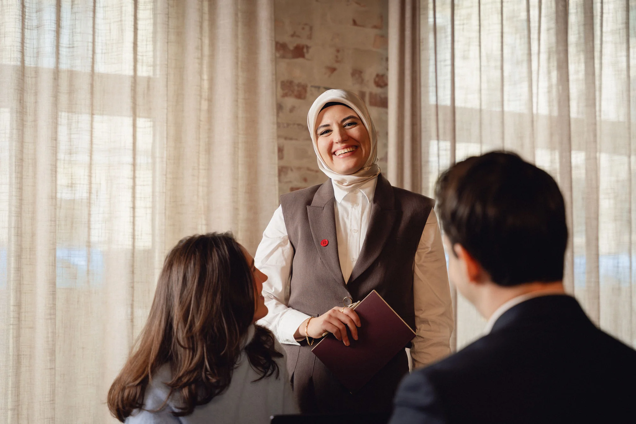 Smilende kvinne i hijab holder notatbok mens hun snakker med to personer i et lyst rom med store gardiner.