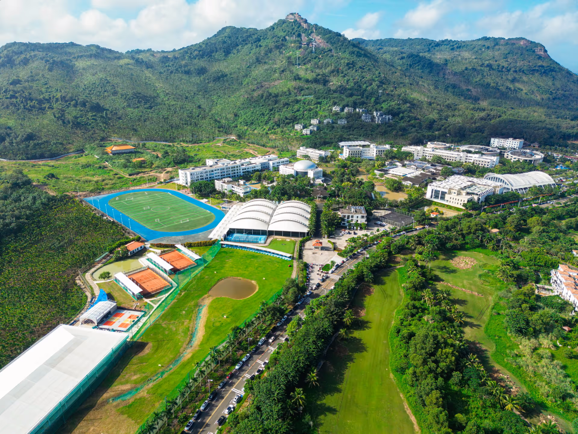 Aerial view of a sports complex with a soccer field, tennis courts, and multiple buildings surrounded by green hills.