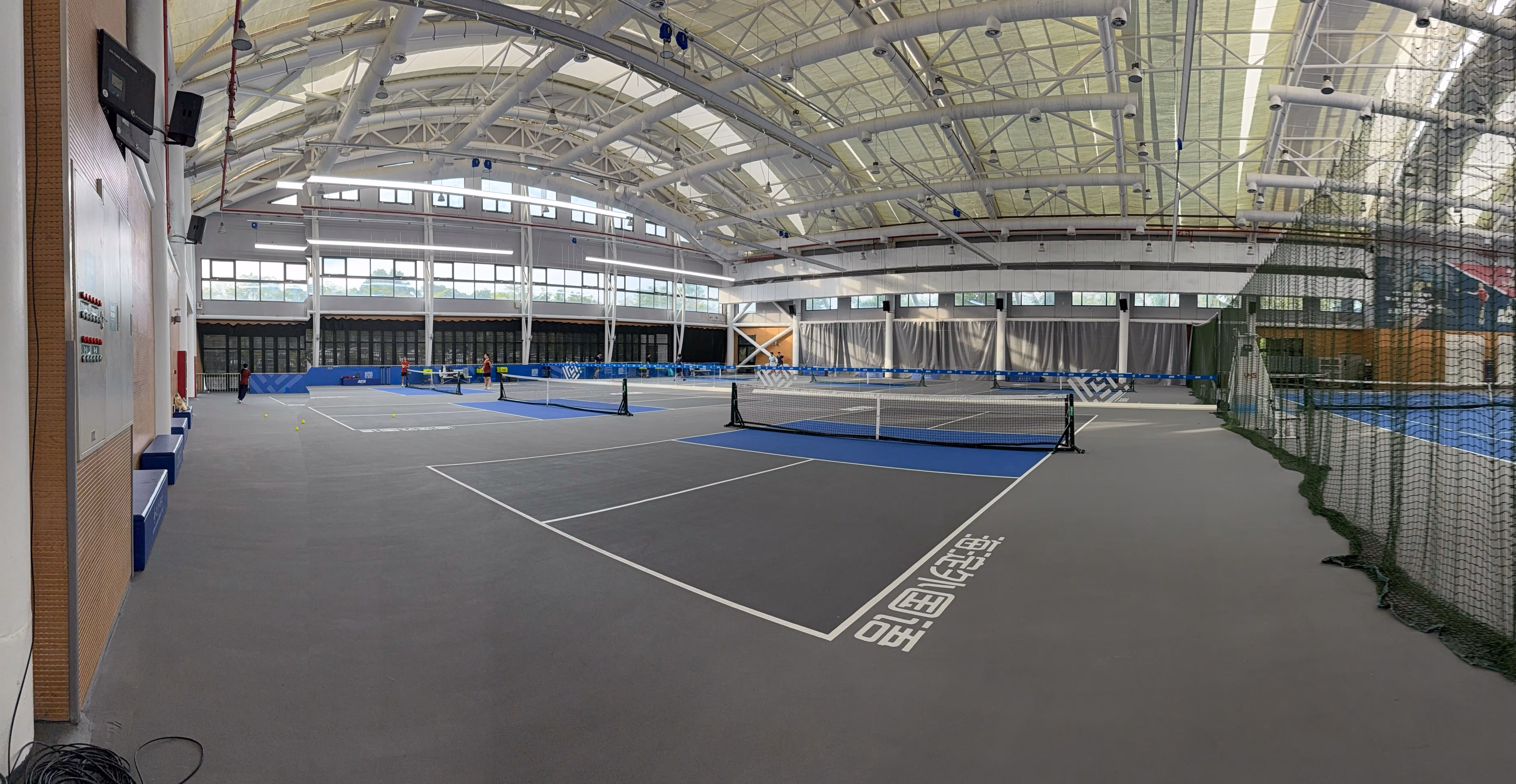Indoor badminton court with multiple nets set up and several people playing, under a high arched ceiling with large windows.