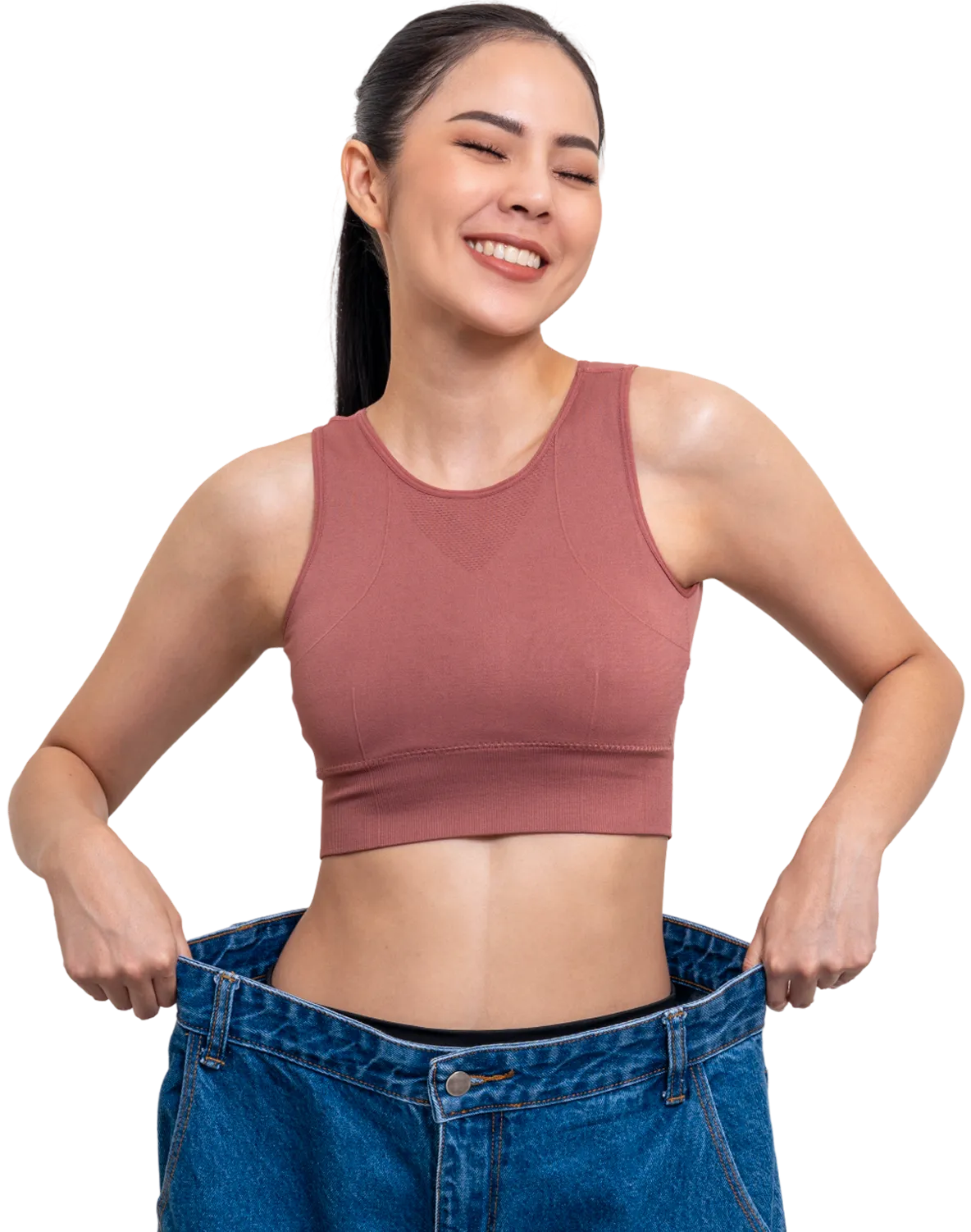 A smiling woman wearing a sleeveless, pink crop top and oversized blue jeans, holding the waistband outward to show weight loss.