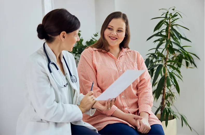 A female doctor in a white coat is holding a paper and conversing with a smiling female patient who is wearing a striped shirt. They are sitting in a well-lit room with green plants in the background.