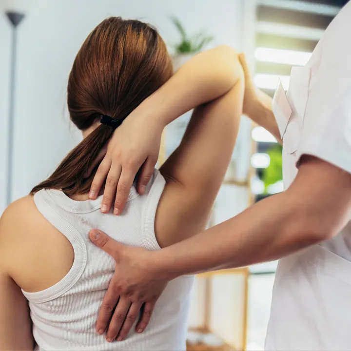 A physical therapist assists a woman stretching her arm and shoulder, with the woman wearing a white tank top and her brown hair tied back.