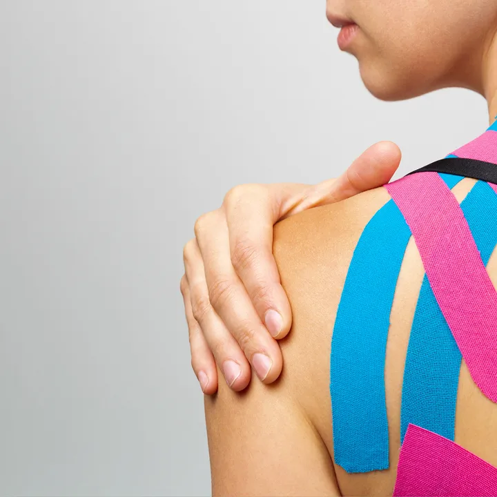 A person with kinesiology tape applied on their shoulder, with three strips—two blue and one pink—extending from the upper back and shoulder toward the upper arm. The person is holding their shoulder with the opposite hand.