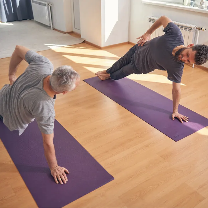 Two men performing side plank exercises on purple yoga mats in a sunlit room with wooden floors.
