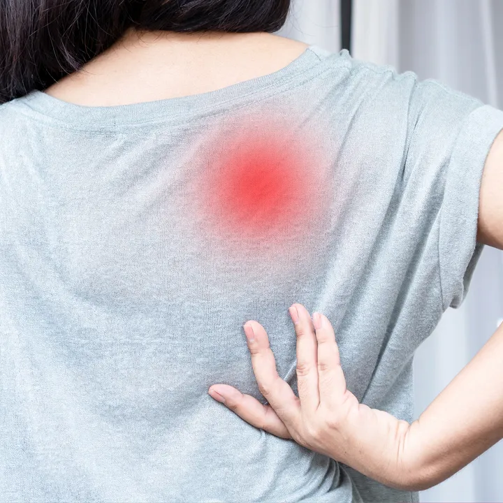 A person in a light gray shirt touching their lower back with one hand, with a red circular highlight on the upper back, indicating pain or discomfort in that area.