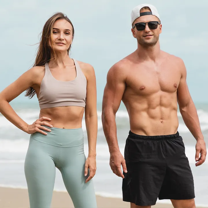 A fit, athletic woman wearing a beige sports bra and light blue leggings stands next to a muscular, shirtless man in black shorts and a white backwards cap, both posing confidently on a beach with the ocean and sky in the background.