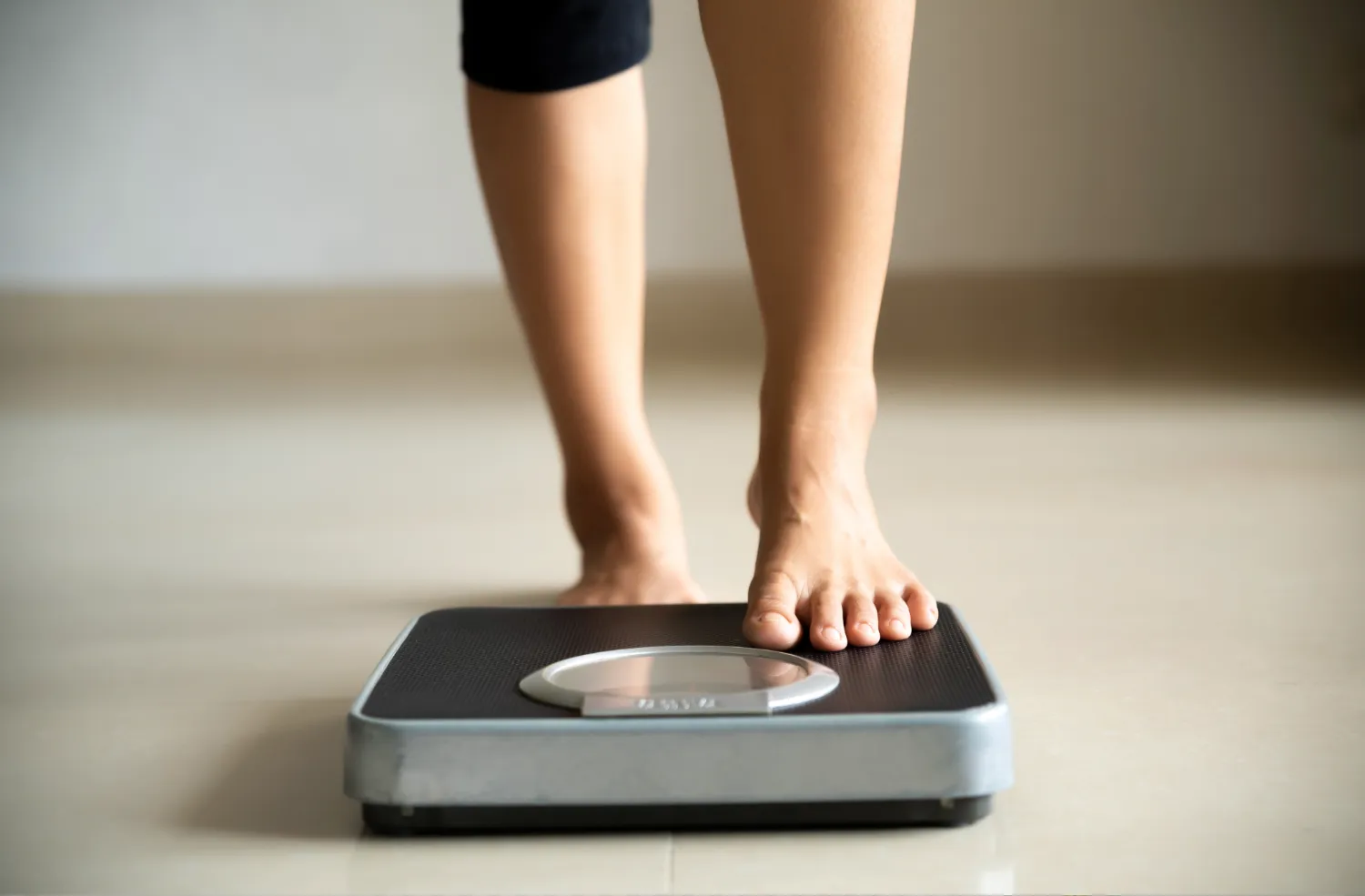 A person stepping onto a digital bathroom scale, positioned on a smooth, beige floor.