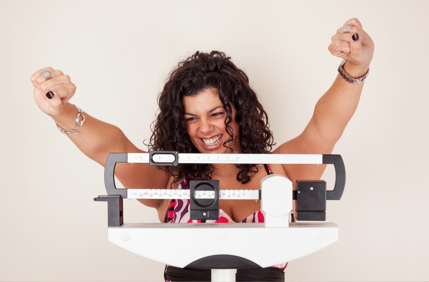 Woman with curly hair celebrating on a mechanical weight scale, looking pleased and raising her fists in triumph.