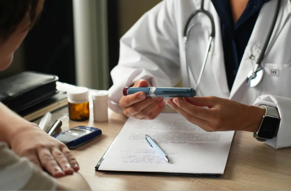 A doctor holding a medication pen while discussing with a patient at a desk, with prescription bottles, a glucometer, and a pen on the table.