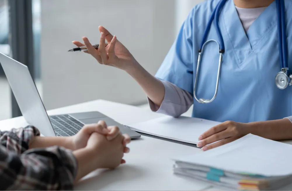 A healthcare professional in blue scrubs and a stethoscope gestures with a pen while talking to a patient across a desk, with a laptop and documents nearby.