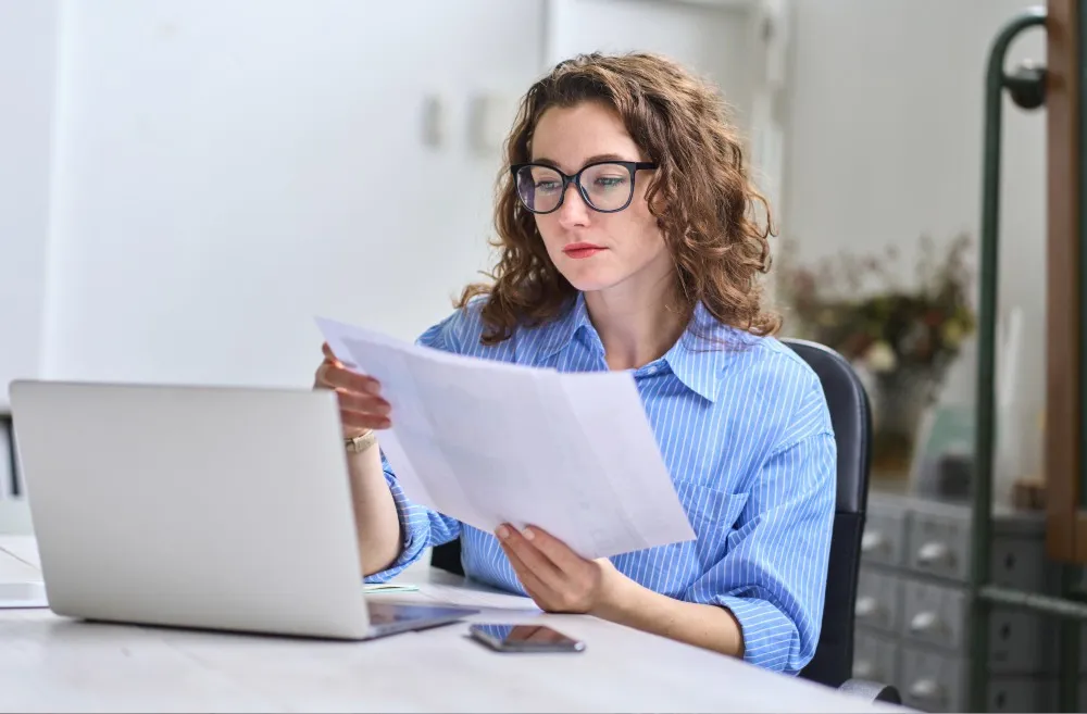 A woman with curly brown hair and glasses is sitting at a desk, wearing a blue pinstripe shirt, reviewing documents in her hand while looking at her laptop. An open smartphone and a pen are on the desk next to her.
