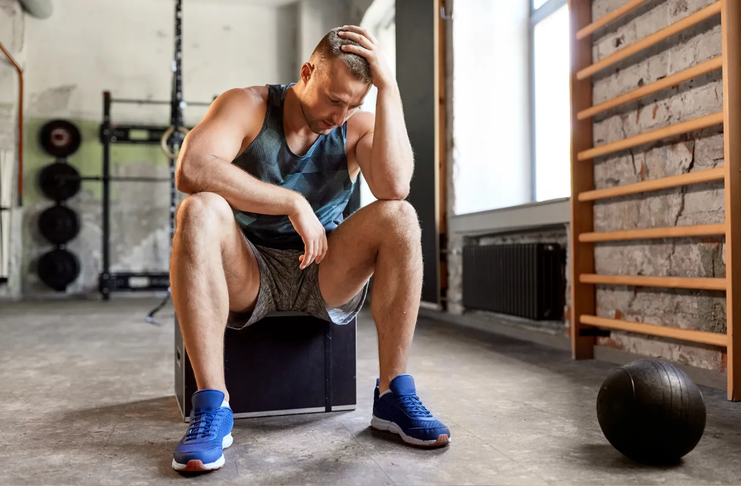 A tired man sitting on a box at the gym, resting with his elbow on his knee and hand on his head, wearing a tank top, shorts, and blue sneakers, with gym equipment in the background.
