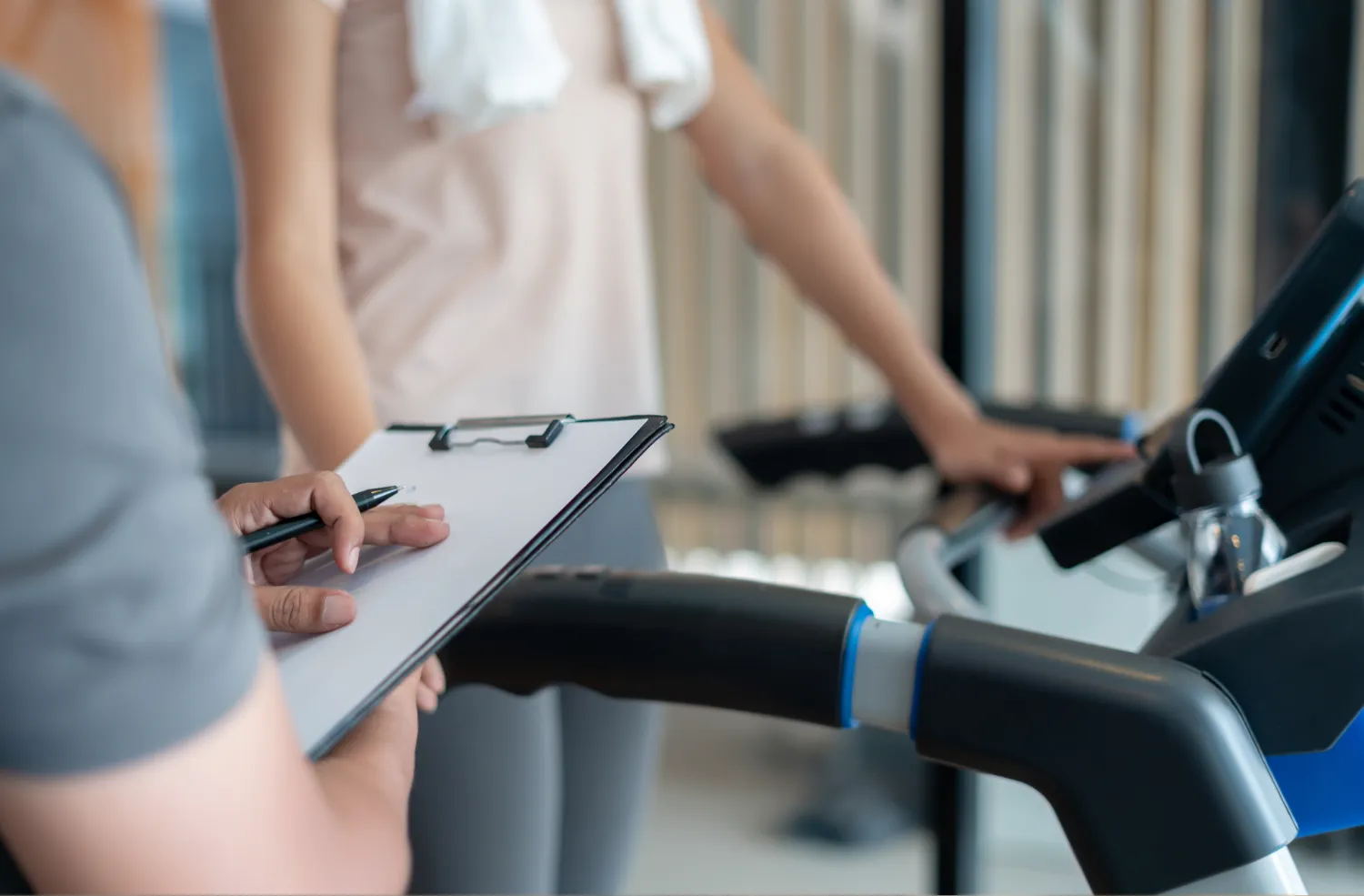 A person in exercise attire is using a treadmill while another individual holds a clipboard, appearing to take notes or track progress.