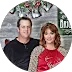 A couple posing in front of a Christmas-themed background with festive decorations. The man is wearing a black shirt and the woman is wearing a red top with light brown lace sleeves.