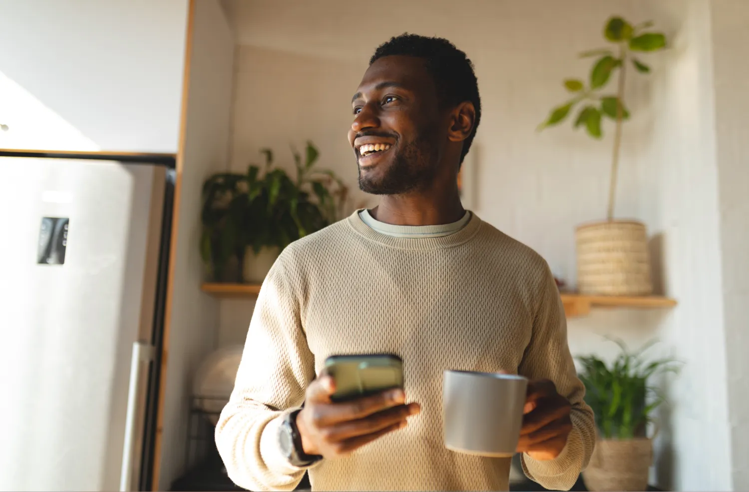 A smiling man holds a smartphone in one hand and a gray mug in the other, standing in a kitchen with plants and wooden shelves in the background.