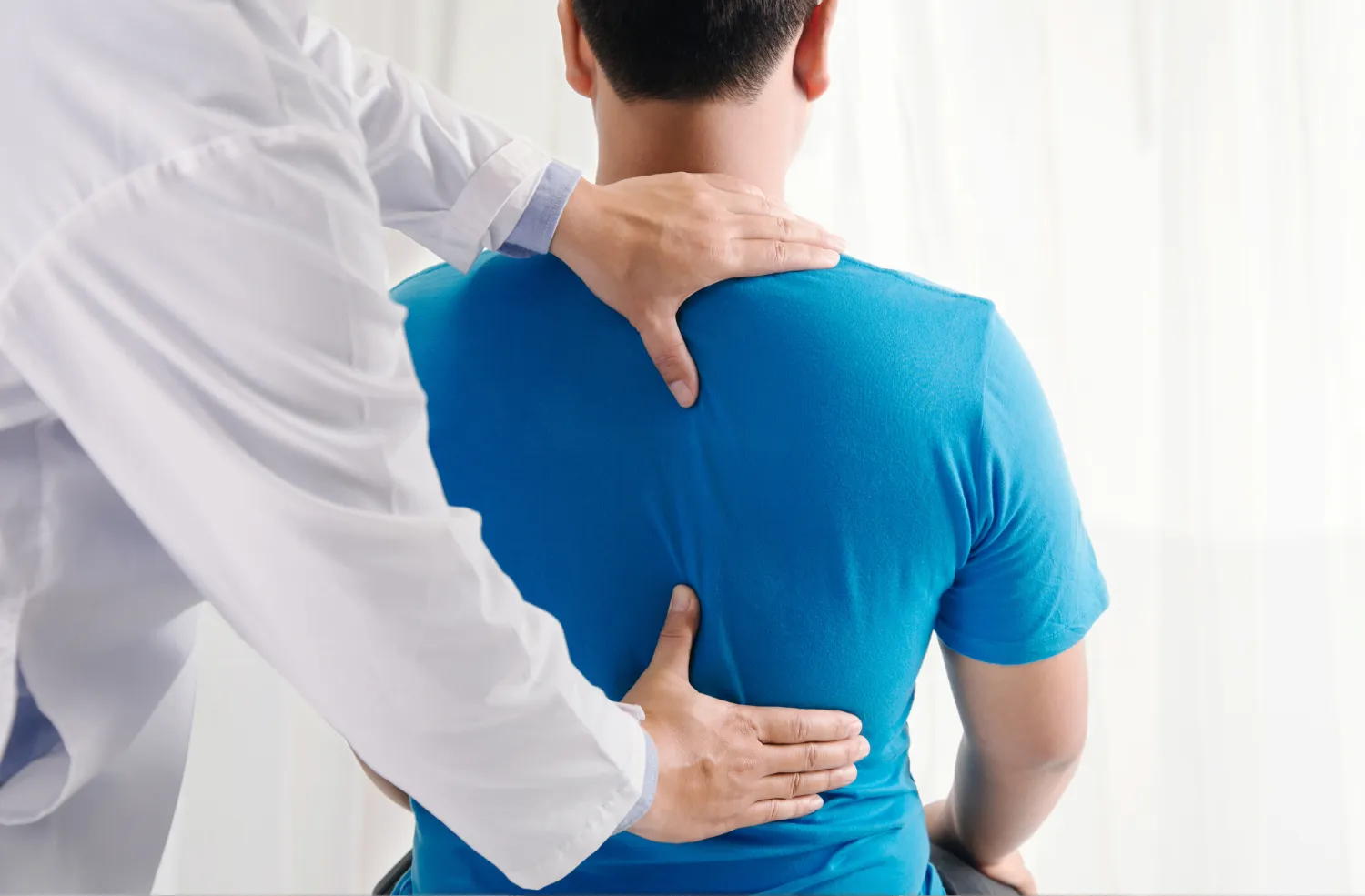 A chiropractor examines a man's back, using one hand to stabilize his shoulder and the other hand to palpate his lower back.