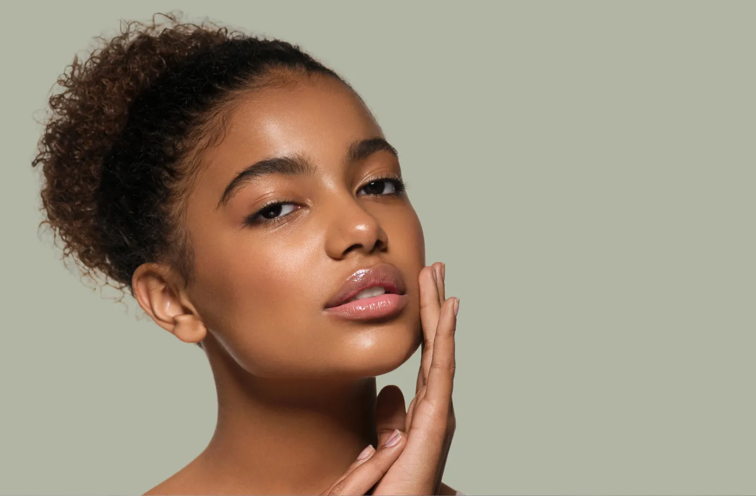 Close-up portrait of a young woman with clear glowing skin and curly hair tied back, gently touching her face.