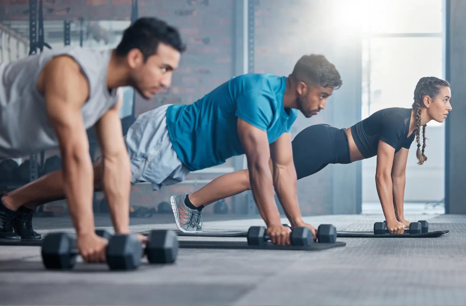 Three people in a gym performing push-ups using dumbbells on exercise mats.