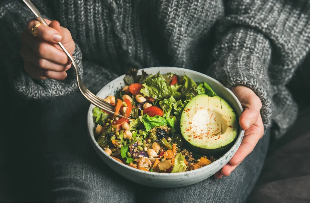 Person in a gray knit sweater holding a bowl of healthy salad with avocado, chickpeas, tomatoes, and greens, eating with a fork.